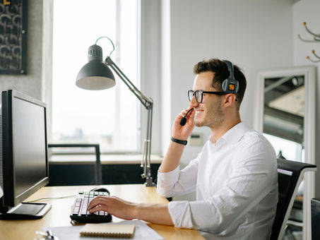 A man working at his computer in an office, wearing a headset and smiling at something on the screen.