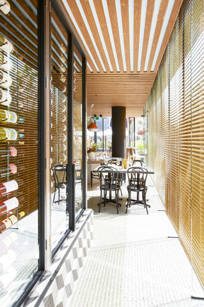 Interior view of a long, bright restaurant verandah featuring dark dining chairs, timber tables, and prominent wood-slat sun louvers.