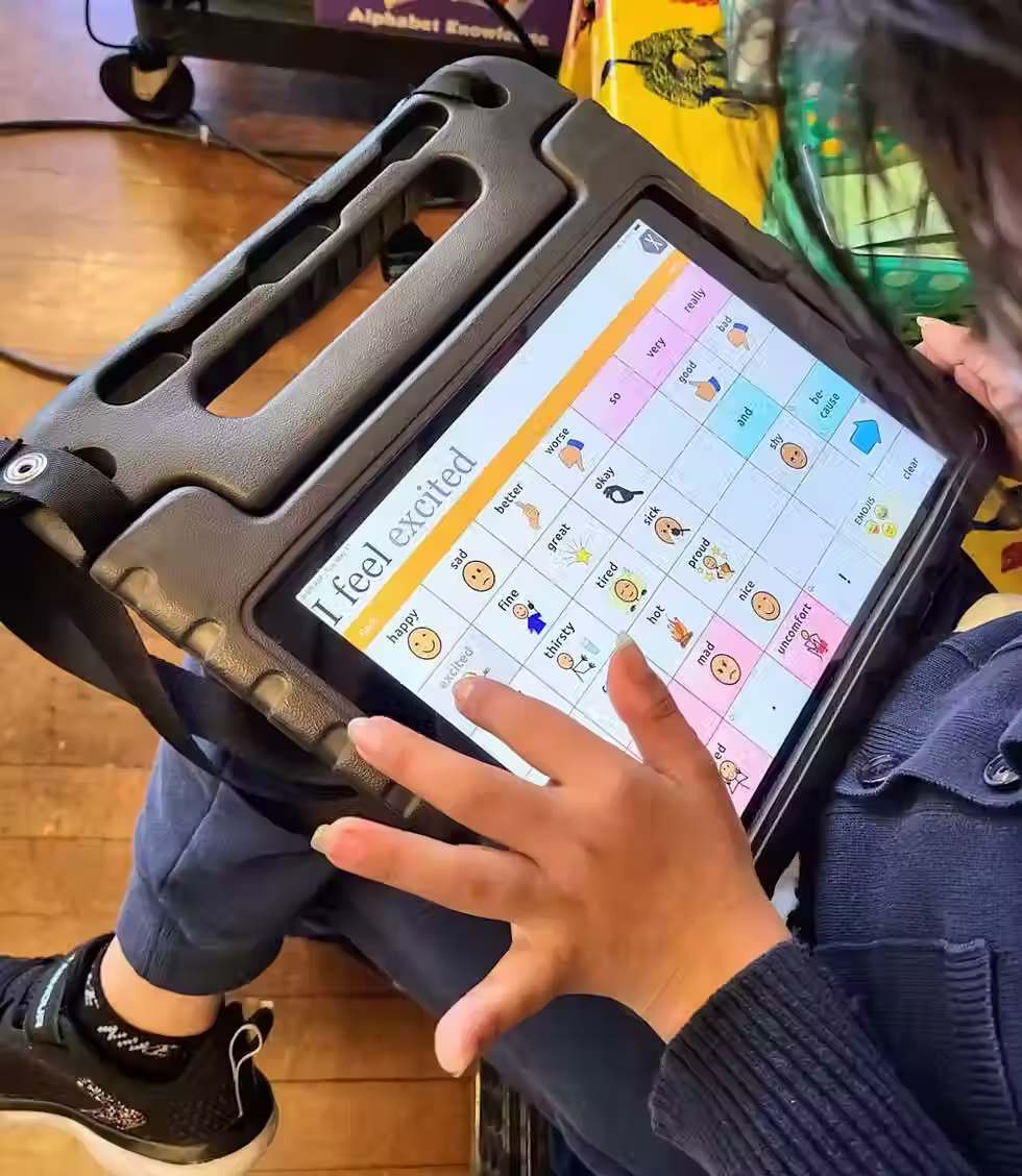 Child using an AAC tablet communication device displaying a symbol-based feelings board, with words and icons to express emotions, during a pediatric speech therapy session.