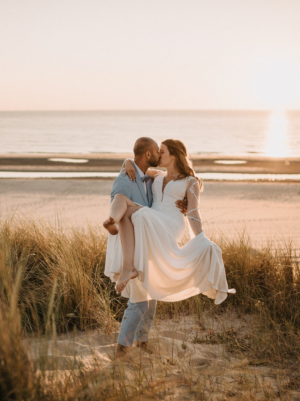 Bruidspaar bij romantische zonsondergang op het strand van Noordwijk — bruidegom tilt bohemian bruid op tijdens strandhuwelijk georganiseerd door wedding planner Anne-Martine Speelman
