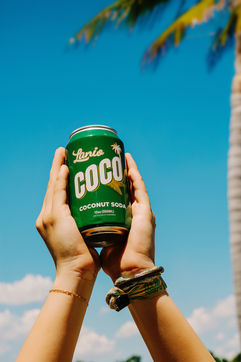 Hands holding a green can of Lani's Coco Coconut Soda against blue sky.