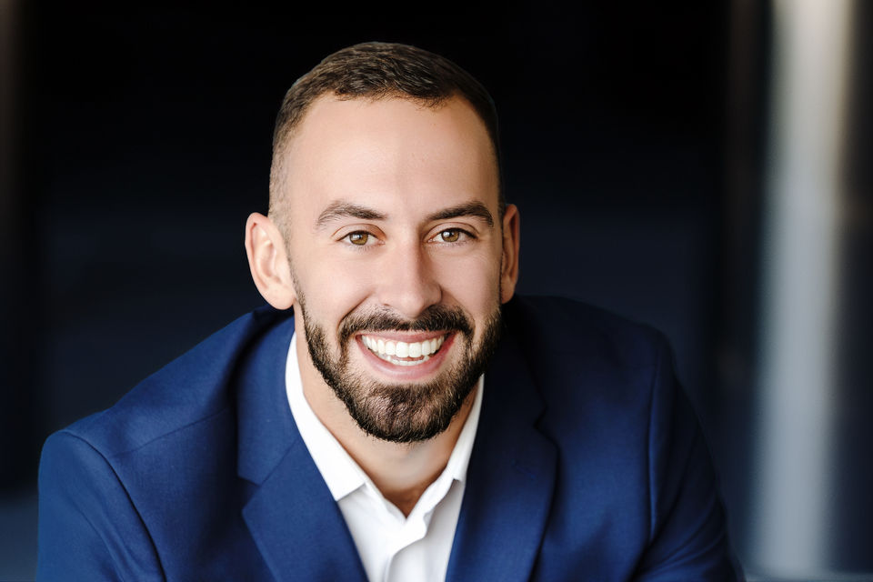 Smiling man in a blue suit, posing for a portrait, with dark background.