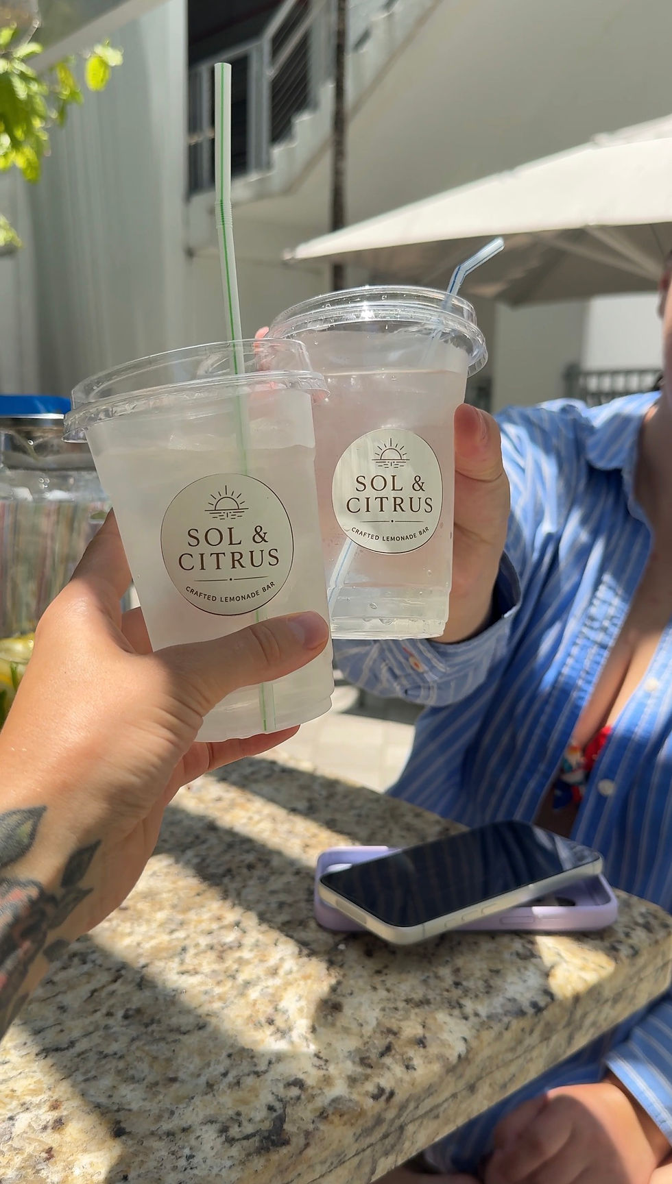 Two branded Sol & Citrus lemonade cups being toasted outdoors on a stone counter, bright sun, lifestyle mobile bar vibe
