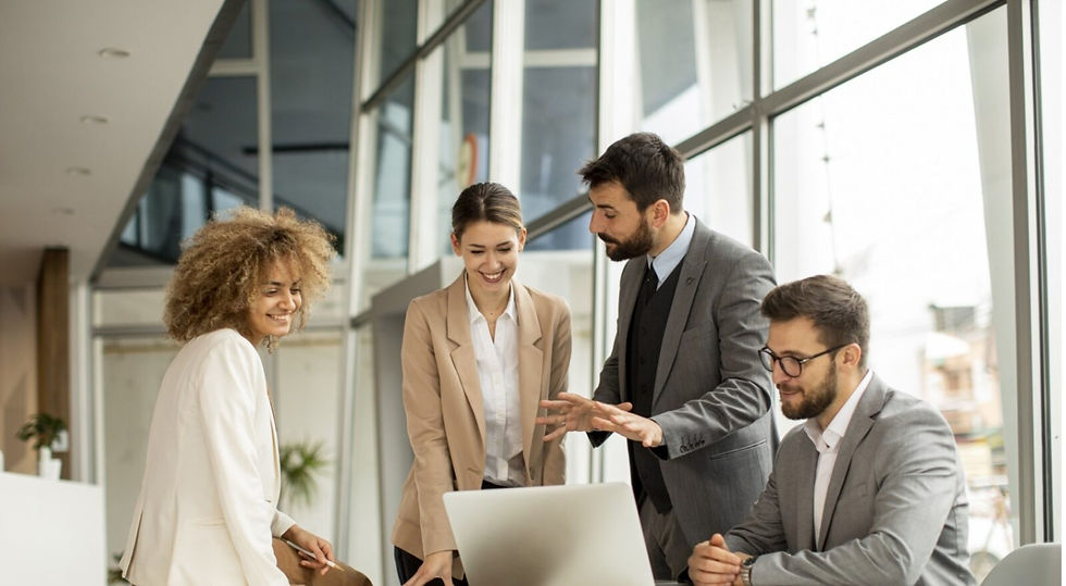 Quatre collègues en costume discutent autour d’un ordinateur portable dans un bureau lumineux, souriants et concentrés.