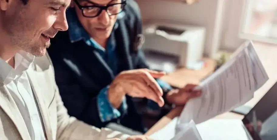 Deux hommes discutent avec des documents à la main dans un bureau. L'homme à lunettes pointe le papier. Atmosphère studieuse et éclairage chaleureux.