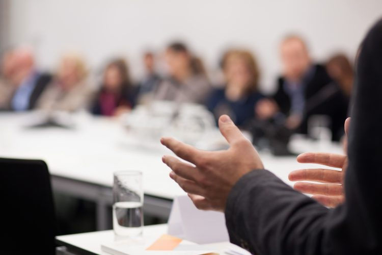 Main à l'avant-plan gesticulant lors d'une réunion dans une salle de conférence. Arrière-plan flou de participants assis à une table.