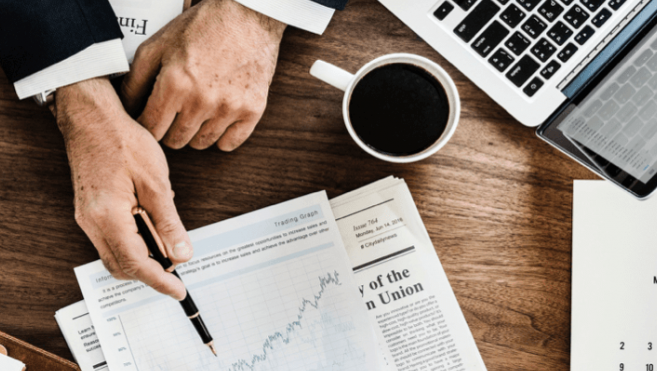Homme en costume pointant un graphique financier sur un bureau en bois, avec un ordinateur portable, café et journaux. Ambiance professionnelle.