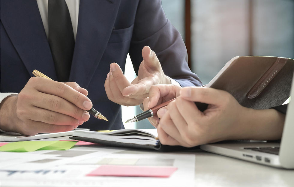 Réunion de travail: deux personnes en costume discutent avec des stylos au-dessus d’un carnet, près d’un ordinateur portable.