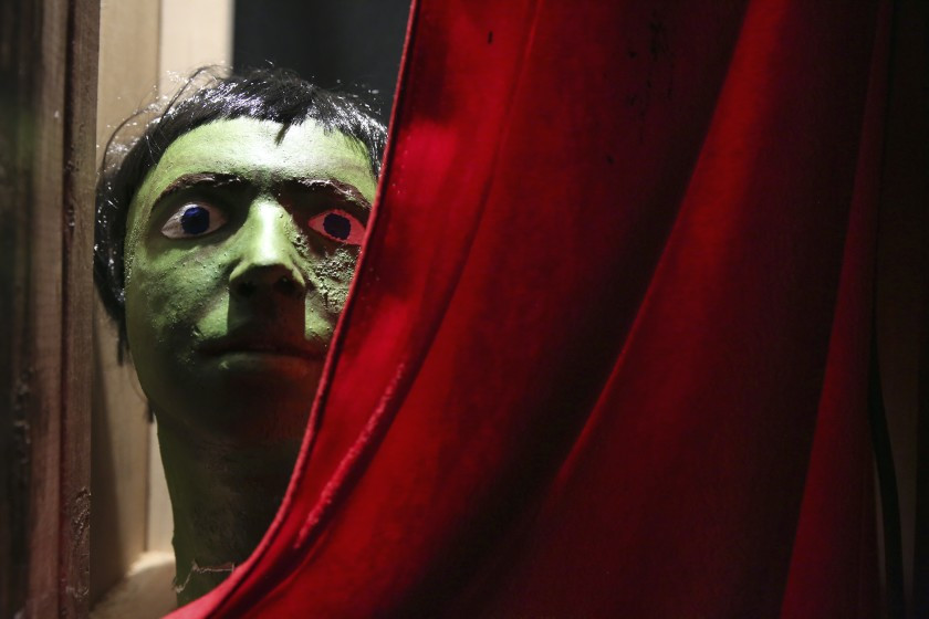 Face in a green cracked mask with painted eyes peering from red curtain