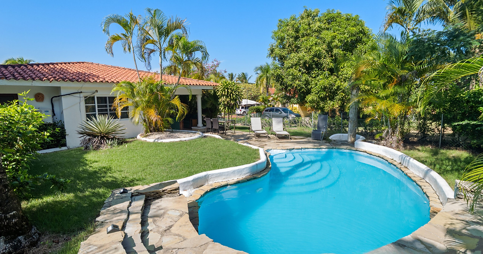 Sunny tropical backyard with a blue curved pool, lounge chairs, palm trees, and a red-tile house under a clear sky.