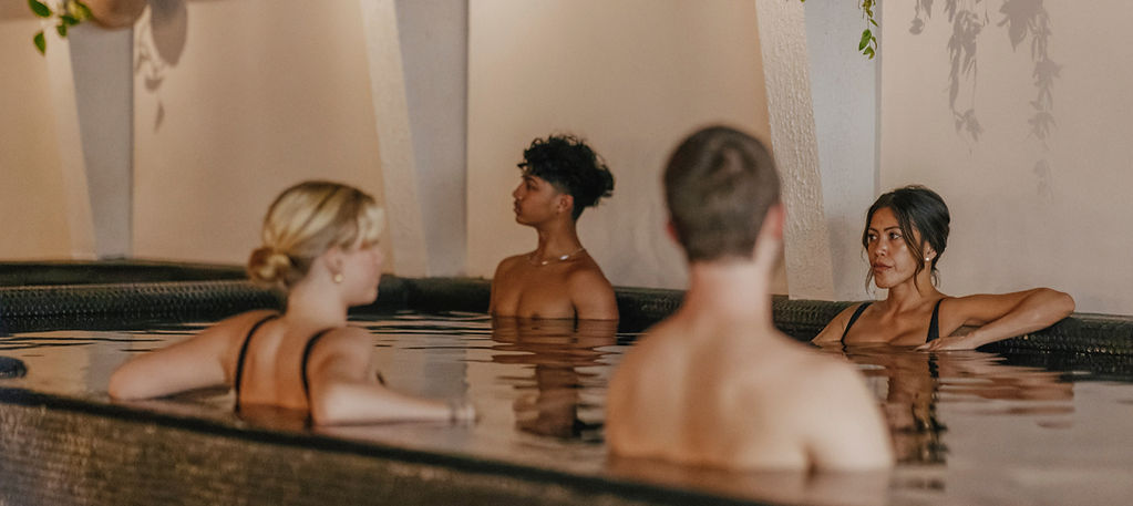 Two people in the foreground facing away, and two in the background. All are soaking in a hot mineral pool. The image has a warm ambiance 