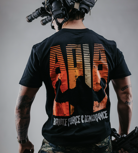 Man in black Ace of Iron Apparel t-shirt holding a tactical rifle, standing against a solid grey background with a bold, mission-ready stance.