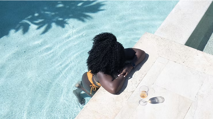 Woman relaxing in a clear blue pool during summer, styled with natural curls, warm sunlight, and a calm luxury aesthetic that captures effortless summer beauty and self-care.