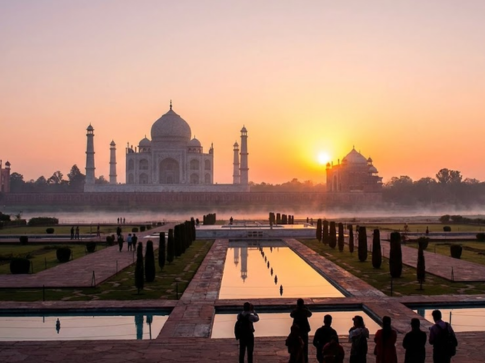 A wide-angle view of the Taj Mahal at sunrise, showing the iconic white marble monument and its four minarets reflected with mirror-like clarity in the central reflecting pool. The sun is a bright orange orb low on the horizon, creating a hazy, golden atmosphere across the grounds. In the foreground, silhouetted visitors stand along the pathways, observing the morning light over the landmark.