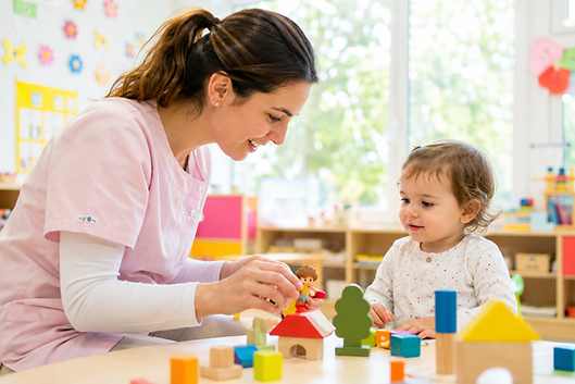 Éducatrice en rose jouant avec un tout-petit à des blocs colorés dans une salle de classe lumineuse et joyeuse
