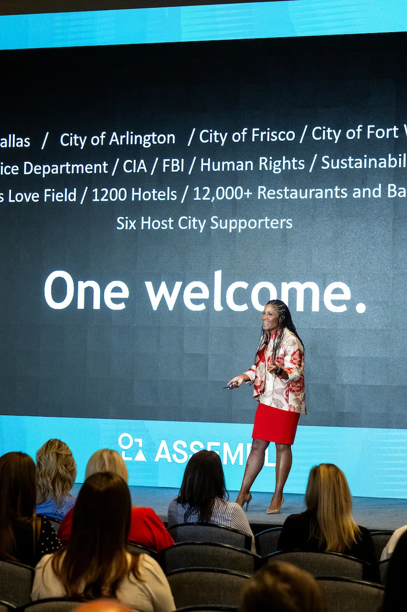 Noelle LeVeaux on stage beside a slide reading One welcome, listing the host city partners