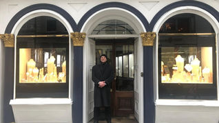 A person in a black coat stands at a jewelry store entrance with arched windows displaying necklaces. The facade is white with blue trim.