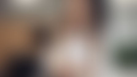 Woman in a white blazer smiles confidently at a sleek office desk. Modern, well-lit space with green plants in the background.