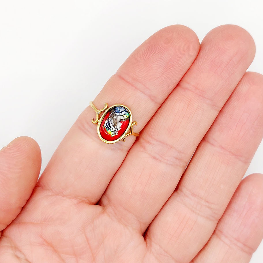 Bague en or avec un portrait peint, portée sur une main, fond blanc.