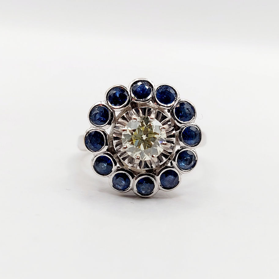 Bague fleur avec diamant central et saphirs bleus, détails de joaillerie sur fond blanc.