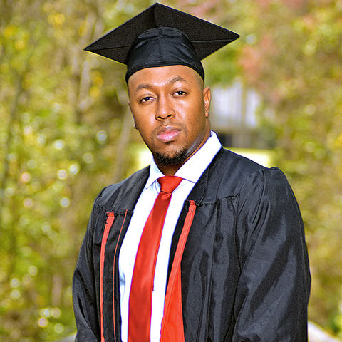 Brandon D. wearing a black graduation cap and gown with a red tie, standing on a wooden path surrounded by trees, looking confidently at the camera.