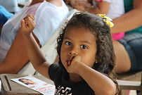 Menina negra, sentada em cadeira em sala de aula olha fixamente para camera.