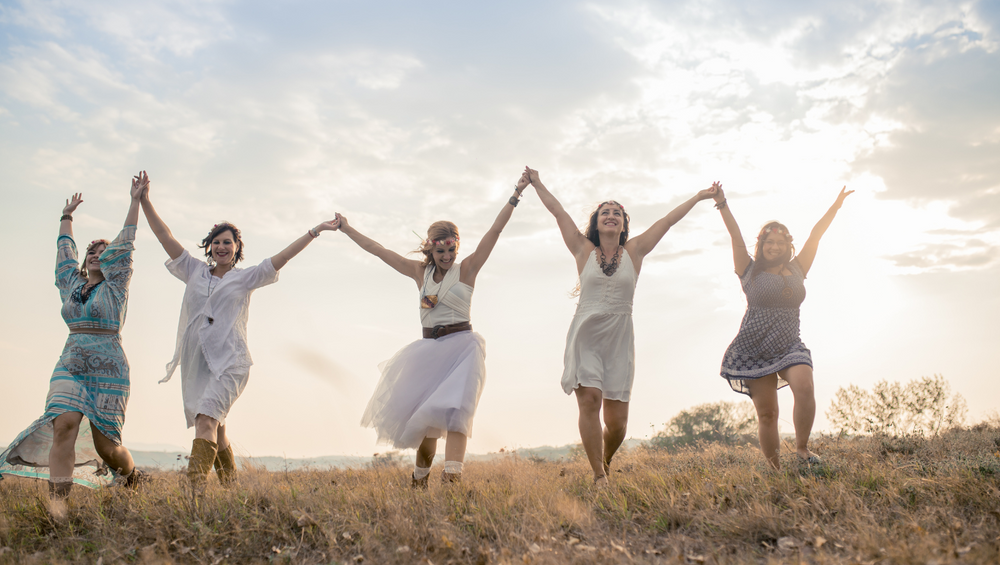 Five women in summer dresses jumping joyfully in unison on what appears to be a grassy hilltop at sunset or sunrise. They are holding hands while jumping, creating a connected line of celebration. Their dresses are in light colors - including white, turquoise patterns, and gray patterns. The scene has a dreamy, carefree quality with the soft lighting from the cloudy sky behind them. The people appear to be having a wonderful time together, with their arms raised and expressions of joy. The natural setting with dried grass and the warm lighting creates a beautiful, candid moment of friendship and happiness.