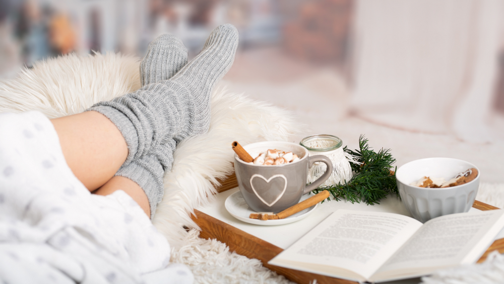 A cozy winter scene showing feet in gray knit socks resting on a white furry blanket, next to a gray mug with a heart design containing hot chocolate topped with marshmallows and a cinnamon stick. An open book lies nearby, alongside some pine branches and a small bowl. The scene suggests comfortable indoor relaxation during cold weather.
