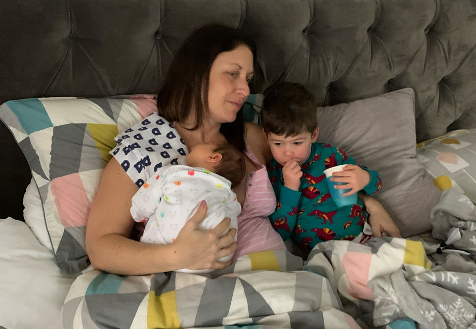 A woman cuddles a baby and a toddler on a bed with geometric-patterned bedding. The toddler drinks from a blue cup, creating a cozy mood.