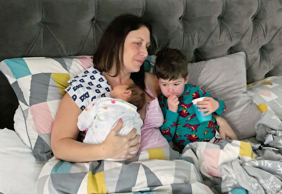 A woman cuddles a baby and child in bed with colorful geometric bedding. The child drinks from a blue cup, creating a cozy, relaxed mood.