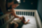 Man smiling at baby in a crib, gently touching the baby's head. Dark room with two small pictures on the wall, cozy and tender mood.