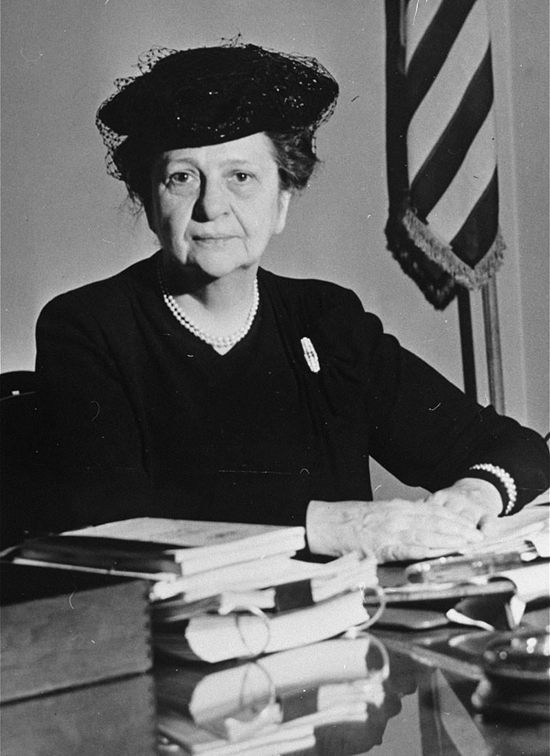 Frances Perkins with a kindly smile in a black hat and dress, sits at her desk among books and documents.