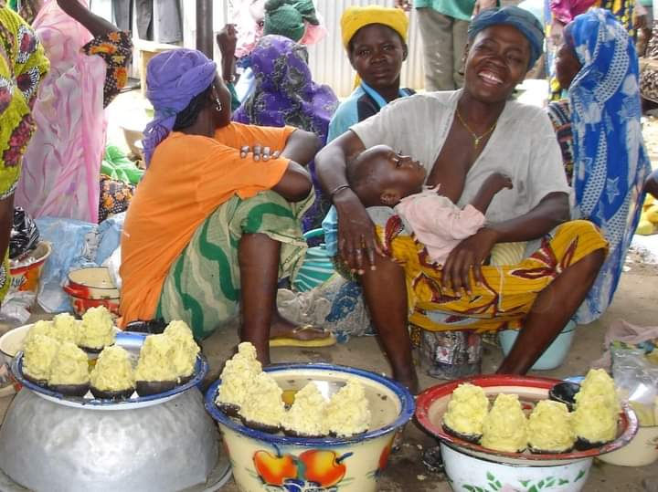 A smiling woman with a child in her lap sits next two other women, with fresh piles of Shea Butter in front of them.