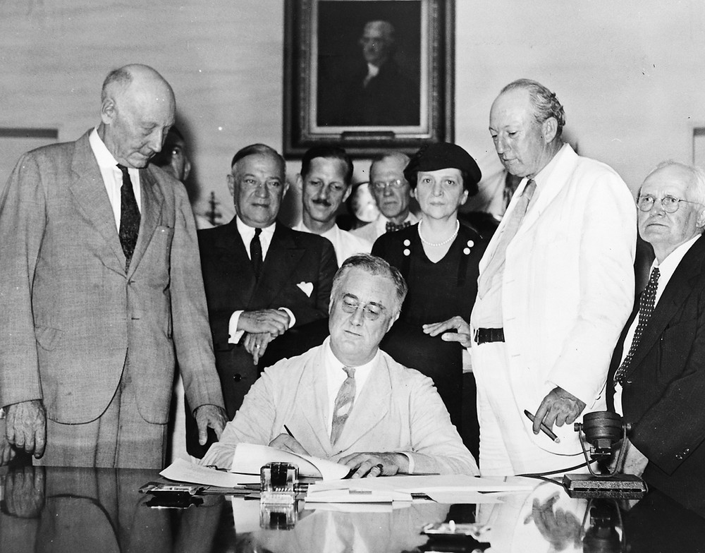 FDR signs the Social Security Act, with lawmakers and cabinet members around him. Frances Perkins stands behind his left shoulder, in a black dress and hat..