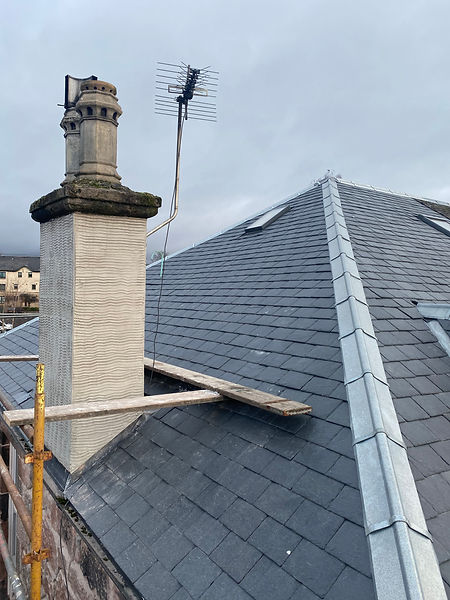 Slate roof with chimney, mounted TV antenna, scaffolding and planks nearby, overcast sky and surrounding buildings visible.