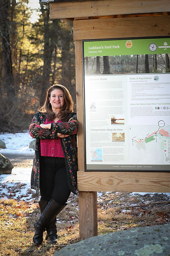 Photo of Lori standing next to sign with arms folded wearing pink shirt and black floral jacket