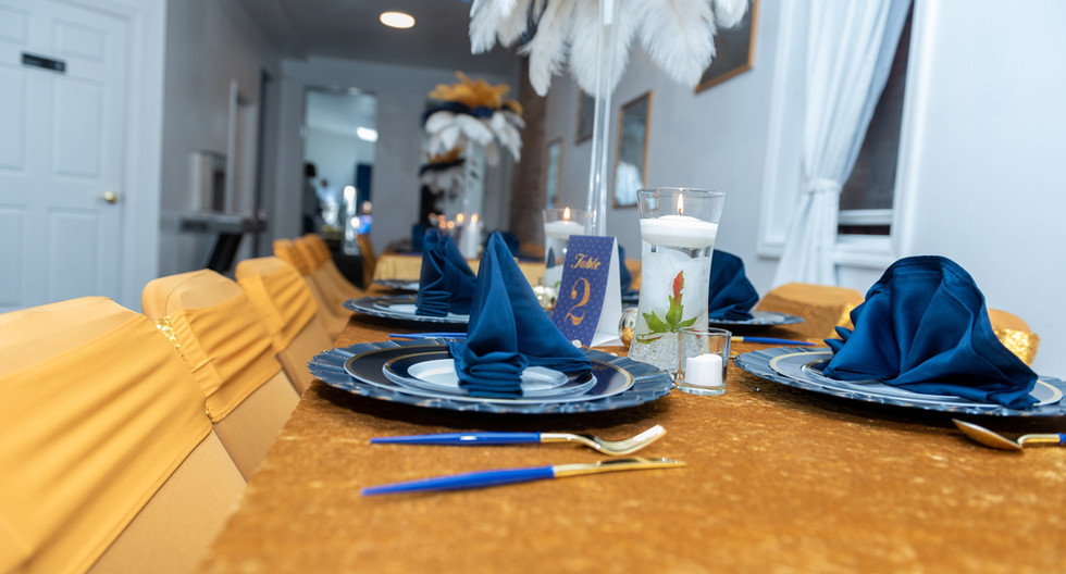 gold, velvet table cloth with gold chair covers and gold sequin chair sashes, with blue, white and gold plates - topped with satin navy blue napkins, chargers, and silverware; completed with empire vases filled with white, blue, and gold ostrich feathers, hurricane glasses filled with water, tea light candles, and acrylic gems.