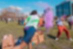 People celebrating outdoors, throwing purple powder in a festive event. Vibrant colors, joyful mood, with buildings in the background.