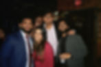 Five people smiling at a formal, wearing nice attire. Background has dark room with string lights and an exit sign. Joyful mood.