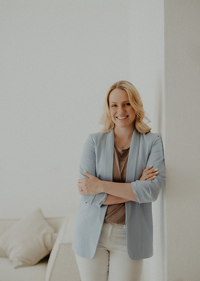 Frau steht vor weißer Wand und lächelt, verschränkte Arme, blondes Haar.