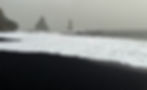 A black sand beach with white waves crashing and two tall, pointed rock formations in the distance under a cloudy sky.