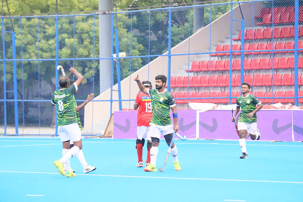 Field hockey players in green jerseys celebrate on a blue pitch, with red stadium seats and a fenced background.