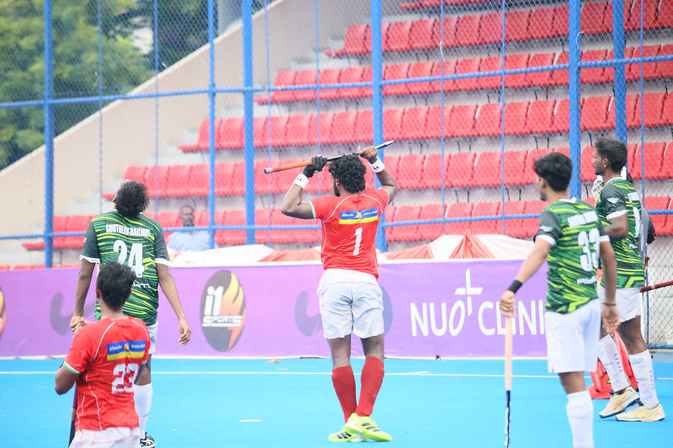 Field hockey players in red and green on a blue turf field; one red player raises his stick, with empty orange stadium seats behind.