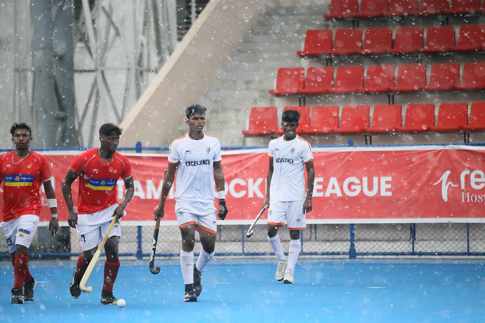 Rainy field hockey match with four players in red and white kits on blue turf, red bleachers and Tamil Hockey League banner.