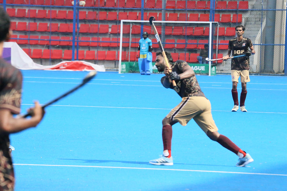 Field hockey players on a blue pitch, one swinging at the ball, with goal and red stadium seats; jerseys read ICF.