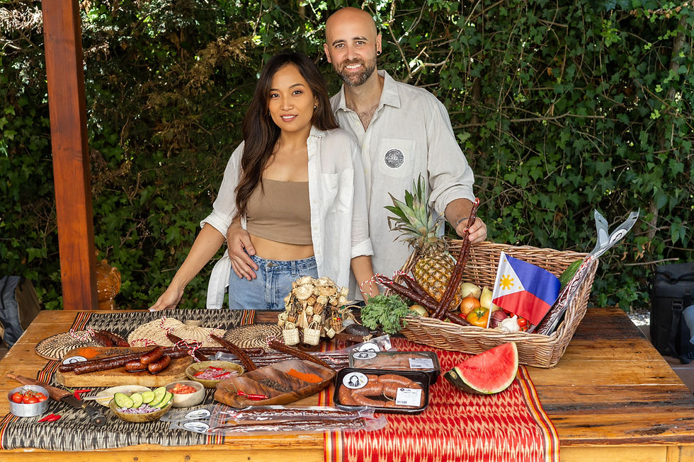 Jelyn Guerra Dimaculangan and her husband pose behind a display of Filipino chorizo, longganisa, fresh produce, and Philippine-inspired food products, representing their family business in Barcelona.