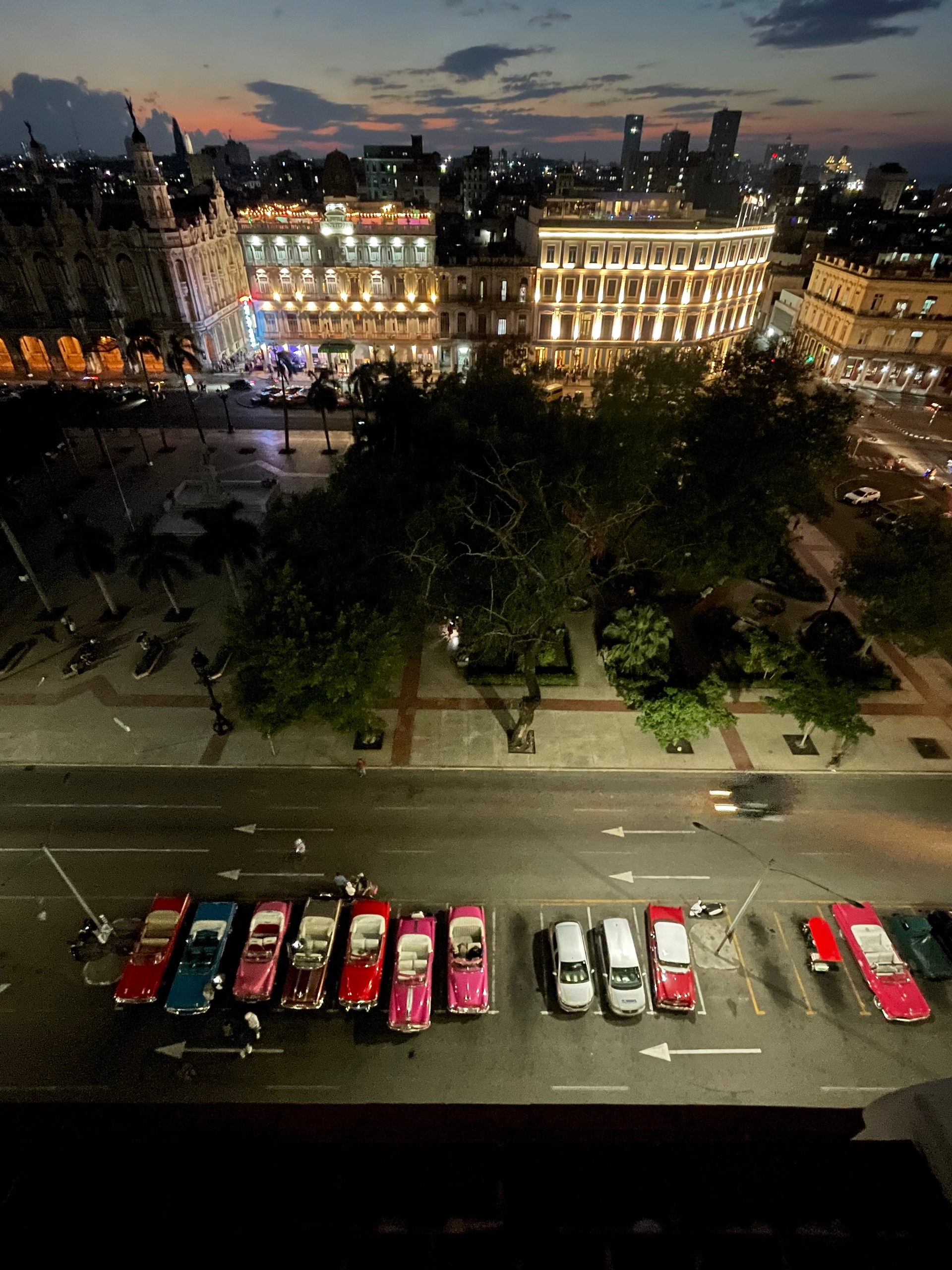 Night view over a brightly illuminated Havana square with classic cars parked along the street