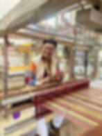 Victor Baguilat Jr., founder and CEO of Kandama, smiles while seated at a traditional wooden handloom inside a weaving workshop. Wearing a jacket made with colorful Indigenous textiles, he demonstrates the weaving process beside threads stretched across the loom.