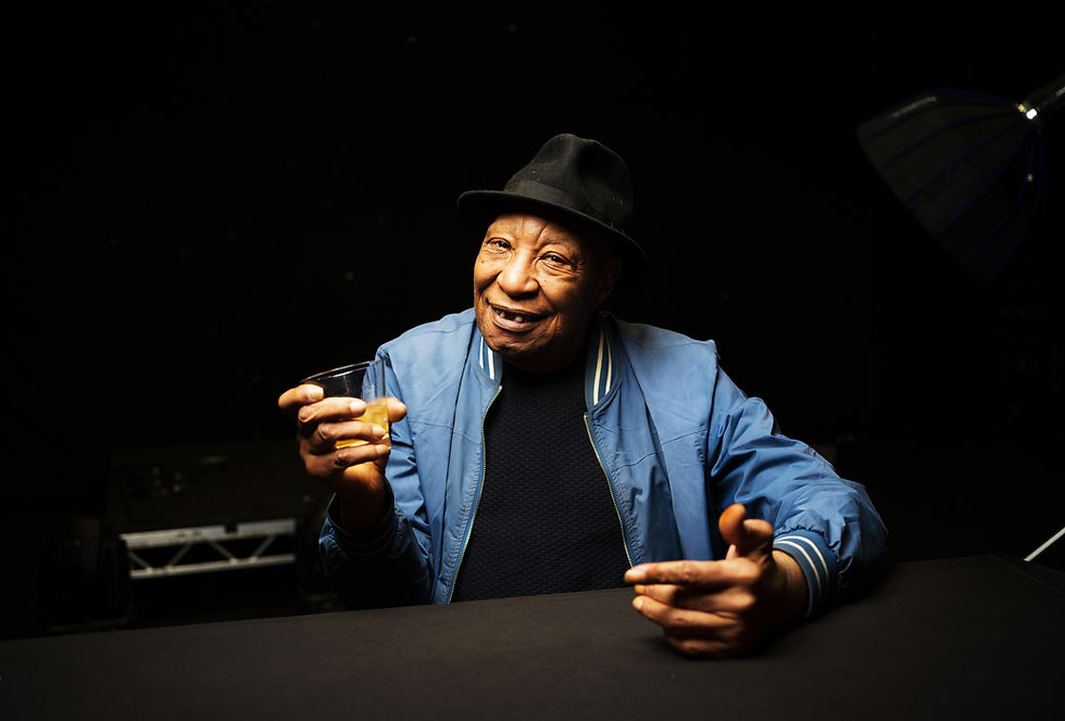 Black man in hos 80s wears a blue jacket and black hat holds a drink in a dimly lit space. The space is a stage - you can see some of the staging rig behind him. The atmosphere is warm and relaxed. the man is smiling gently.