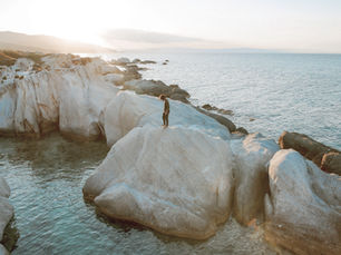 Person standing alone on large coastal rocks by the ocean at sunset, representing implicit memory and strong emotional responses without a clear narrative.